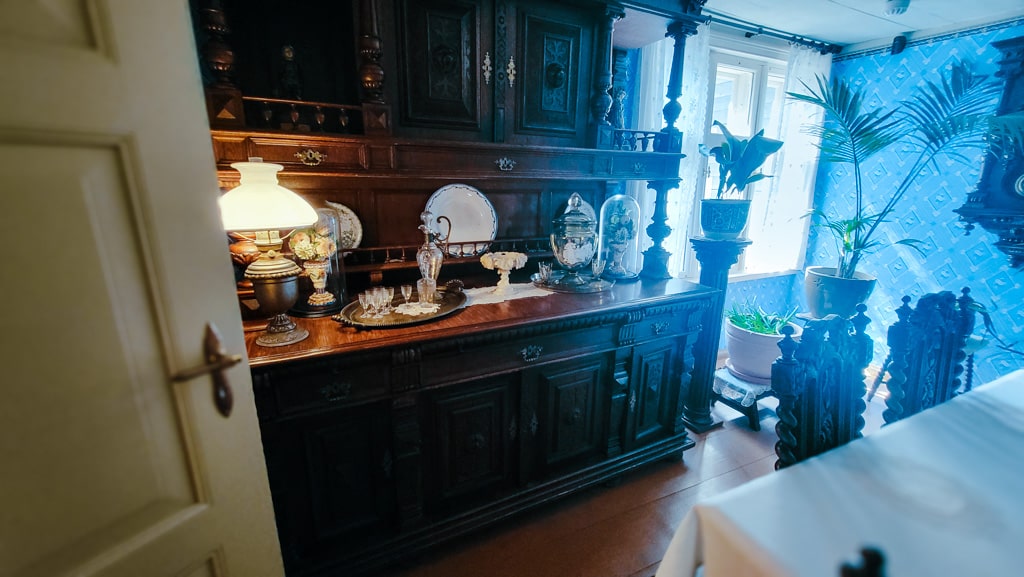 Antique furniture and decor in a historic house museum in Liepaja, Latvia. Features include a dark wood sideboard, vintage lamp, and blue patterned wallpaper.