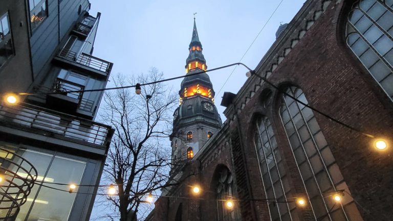 An upward view of St. Peter's Church in Riga, featuring its iconic spire illuminated in warm light against the evening sky. Strings of hanging lights add a cozy ambiance, contrasting with the brick architecture and modern adjacent buildings.