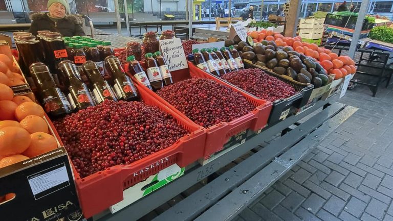 A vibrant fruit and berry stall at Riga Central Market showcasing crates filled with fresh cranberries, bottles of cranberry juice, and sauces. The stall also features avocados, tomatoes, and oranges arranged neatly, with a sign displaying prices in euros.