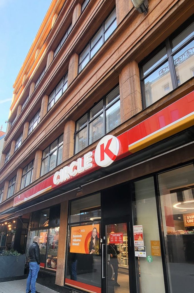 A street-level view of a Circle K convenience store in a multi-story building with large windows. The store features a bold red and white Circle K logo above the entrance, with a man standing outside looking at the window display.