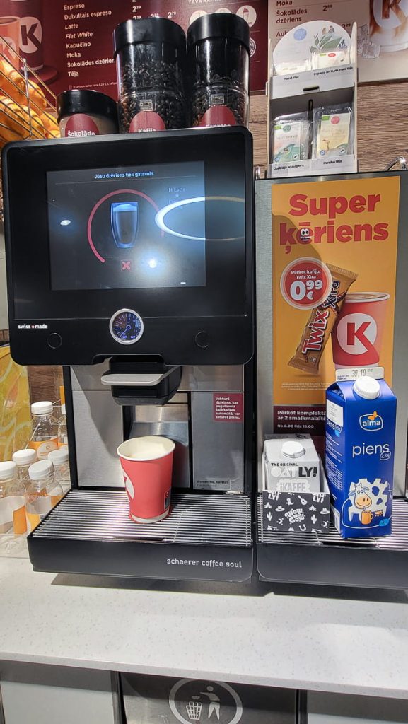 Close-up of a modern coffee machine dispensing a drink into a red Circle K cup, with nearby milk cartons and a promotional sign offering Twix chocolate bars for €0.99.