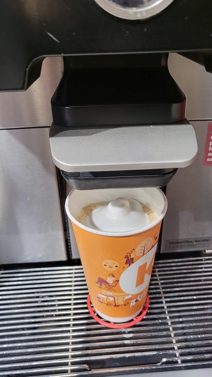 A Circle K coffee machine pouring a frothy coffee drink into an orange cup decorated with cheerful illustrations and the Circle K logo.