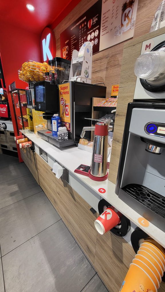 A wide-angle view of Circle K’s self-service coffee station, showcasing multiple coffee machines, syrup dispensers, stacked cups, and promotional signs advertising coffee deals.