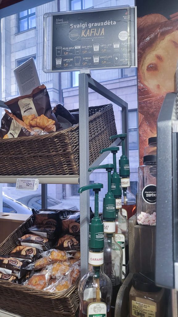 A display of "Teisseire" coffee syrups in caramel and chocolate flavors with green pump dispensers, placed beside wicker baskets filled with assorted packaged pastries.