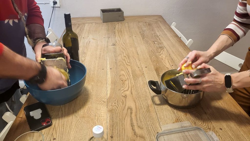 Two people standing at a wooden table, grating food into separate bowls. One person is using a box grater over a blue bowl, while the other is zesting a lemon into a pot. A bottle of wine and various kitchen items are scattered on the table.