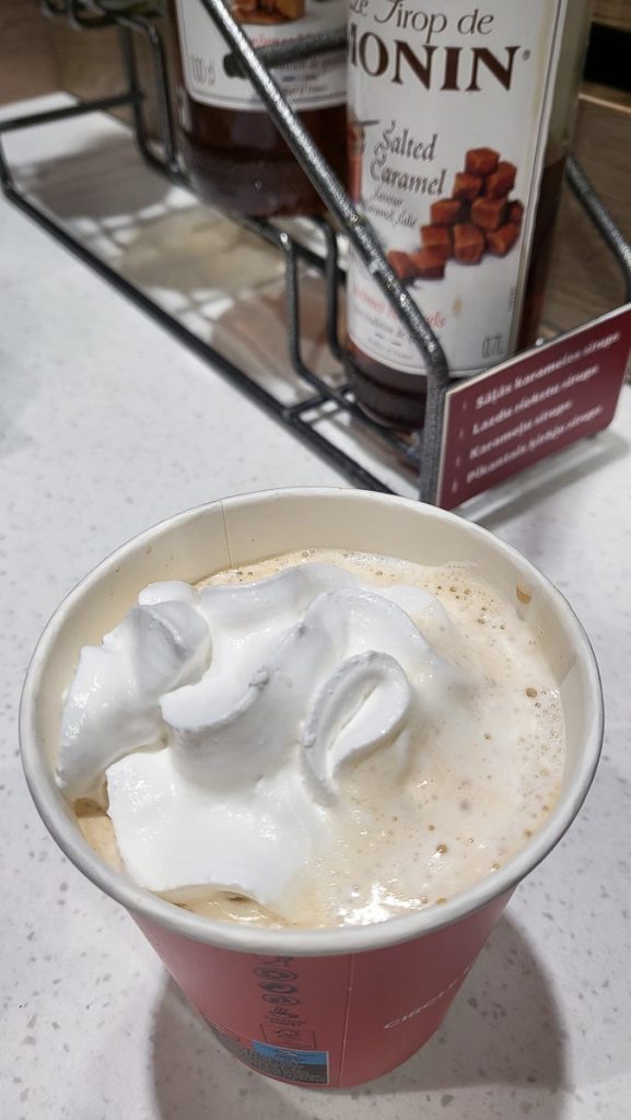 A close-up of a red Circle K coffee cup filled with a frothy coffee drink, topped generously with whipped cream. Bottles of salted caramel syrup are visible in the background.