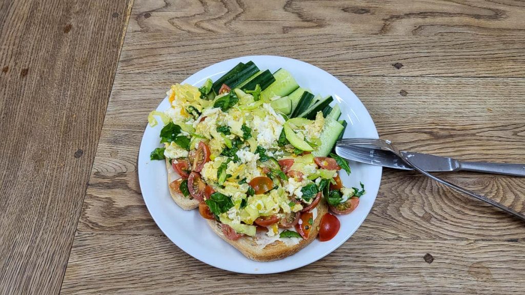 A white plate with scrambled eggs mixed with cherry tomatoes, spinach, and cheese, served on toasted bread. Fresh cucumber slices are arranged on the side. The dish is placed on a wooden table with a knife and fork beside it.