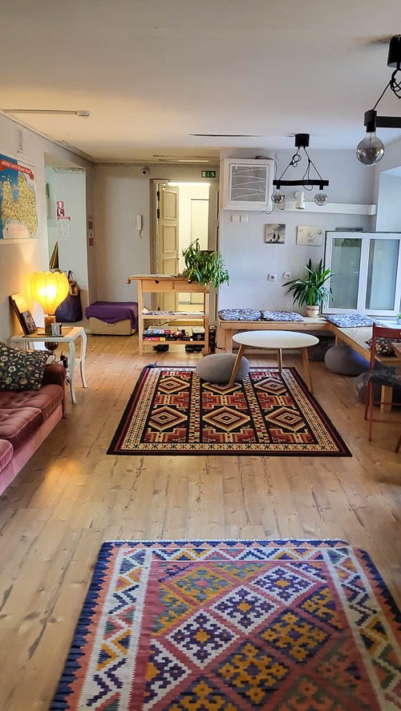 A warm and inviting common area in a hostel with colorful patterned rugs, wooden furniture, soft cushions, and houseplants. The room has an open design with natural light coming in through the windows.