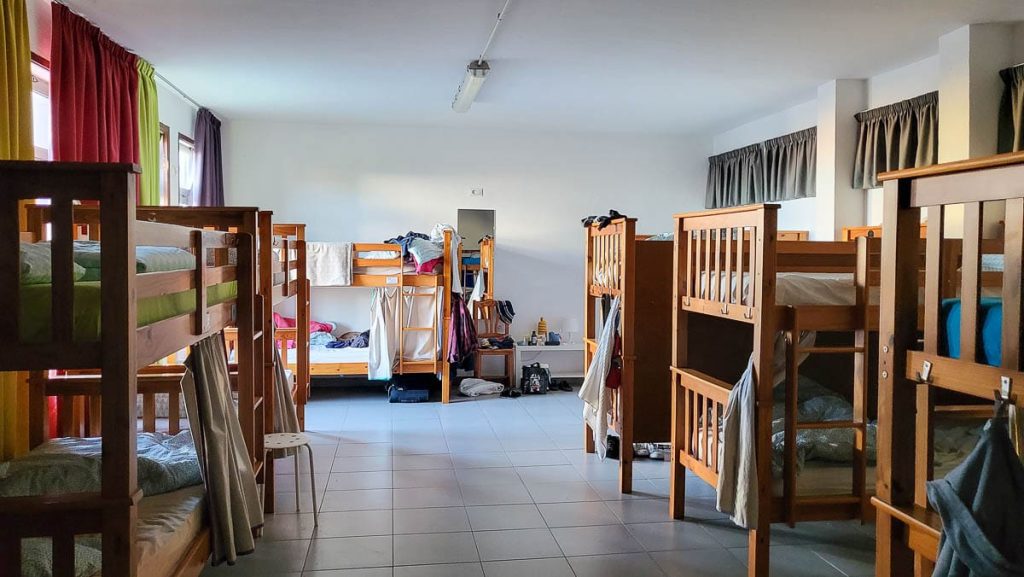 A spacious hostel dormitory with multiple wooden bunk beds arranged in rows. Personal belongings, clothes, and bags are scattered around, giving the room a lived-in feel. Sunlight streams through the windows, illuminating the simple tiled floor.