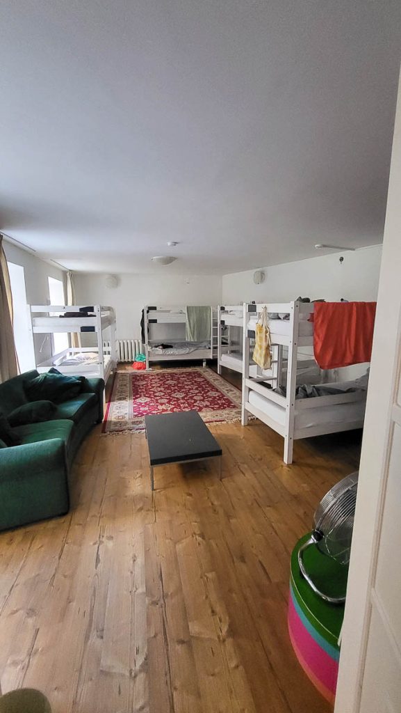 A dormitory-style hostel room with multiple white bunk beds, a red patterned rug, a green couch, and personal belongings scattered around. The room appears well-lit with natural light.