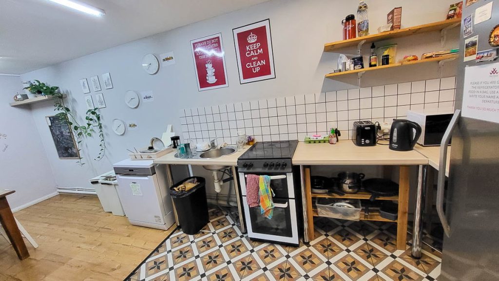 A clean and functional hostel kitchen with white-tiled walls, a stovetop, an oven, a dishwasher, and a sink. The shelves are stocked with cooking essentials, including spices and kitchenware. A "Keep Calm and Clean Up" sign hangs on the wall.