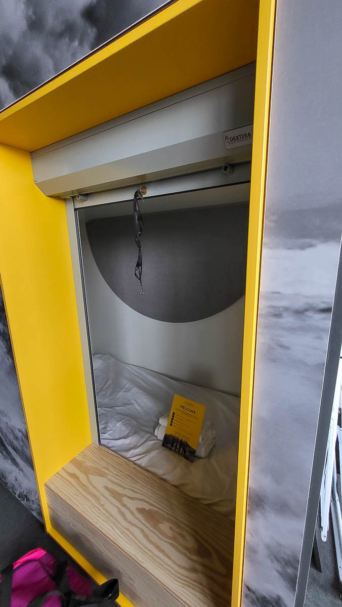 A sleek and modern sleeping pod in a hostel, framed by a yellow border. Inside, there is a bed with white sheets and a welcome note placed on top. A pull-down shade provides privacy.