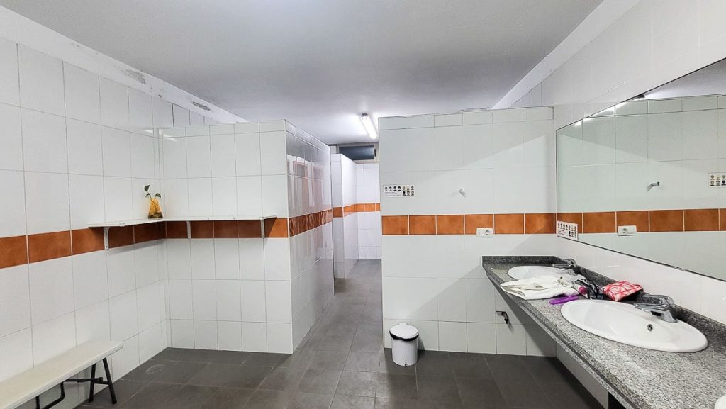 A spacious shared bathroom with white tiled walls, accented by an orange stripe. It includes multiple shower stalls, a large mirror over a dual-sink countertop, and a few personal toiletries on the counter. A small sign with symbols is posted on the wall.