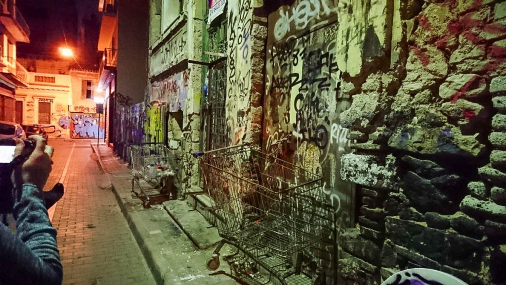 A narrow urban alleyway at night, covered in graffiti on the walls and doors. Several abandoned shopping carts are lined up against the wall. The dim street lighting casts a moody glow over the rough, textured surfaces.