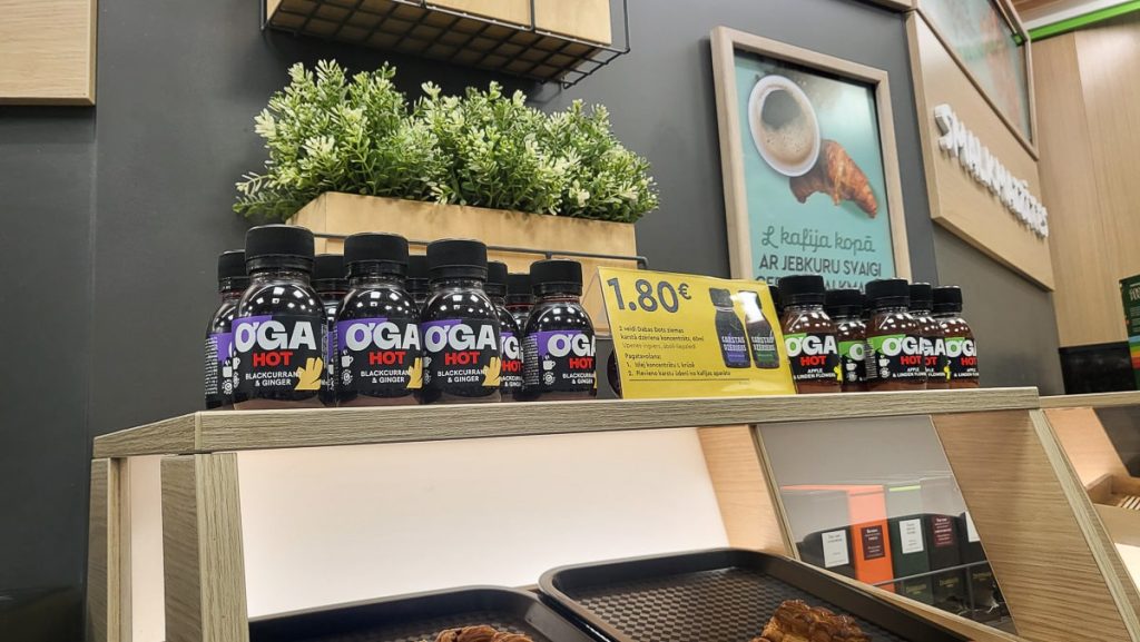 A countertop display of Oga Hot blackcurrant and ginger drinks in small black bottles, priced at €1.80. The display is complemented by a potted plant and a coffee and croissant poster in the background.