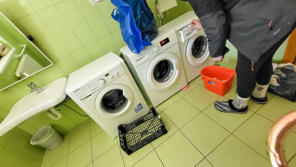 A hostel laundry room with three front-loading washing machines lined up against a lime green tiled wall. A person in warm socks and slippers is adjusting a machine, with laundry baskets and detergent scattered nearby. A small sink and mirror are visible to the left.