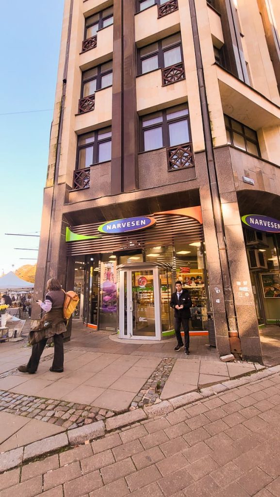 The entrance of a Narvesen convenience store in a multi-story building with dark stone and beige walls. The store features a blue and green Narvesen sign, with people entering and exiting.