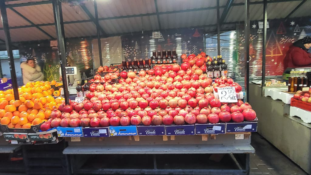 A colorful pomegranate display at Riga Central Market, with rows of pomegranates stacked neatly and a price sign reading "2.90 EUR/kg." Oranges and juice bottles are also visible on the sides, with festive decorations in the background.