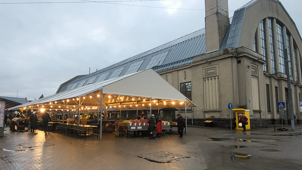 An outdoor view of Riga Central Market, featuring a covered market area illuminated with warm string lights. The historic market building with its arched windows stands in the background, while people shop at various stalls under the canopy.