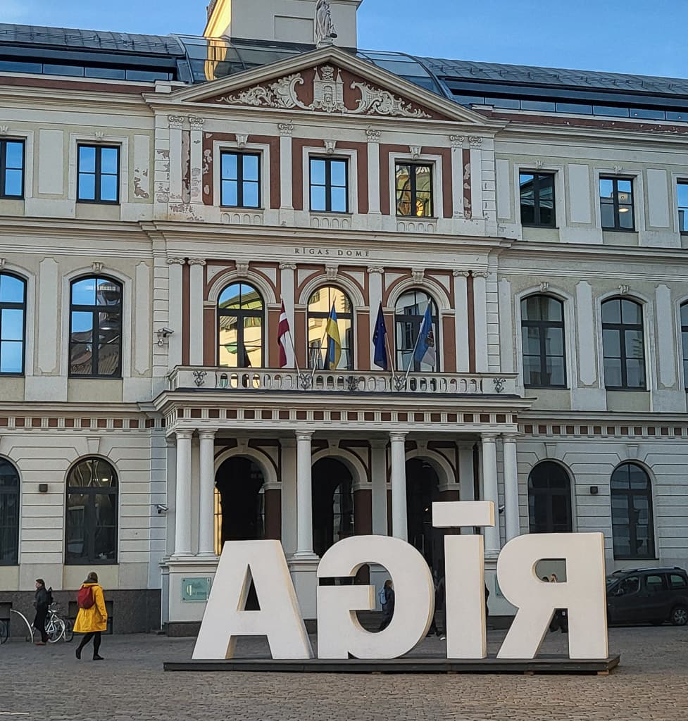 Riian kaupungintalo Vanhassakaupungissa, jonka edessä on suuri "RIGA"-kirjainveistos takaapäin kuvattuna. Keltatakkinen henkilö kävelee torilla, tuoden eloa maisemaan.