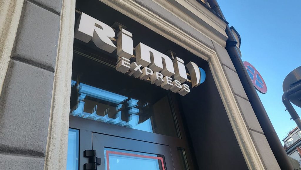 Close-up of the "Rimi Express" sign above a store entrance on a beige building exterior. The sky is clear and blue with a visible no-parking sign nearby.