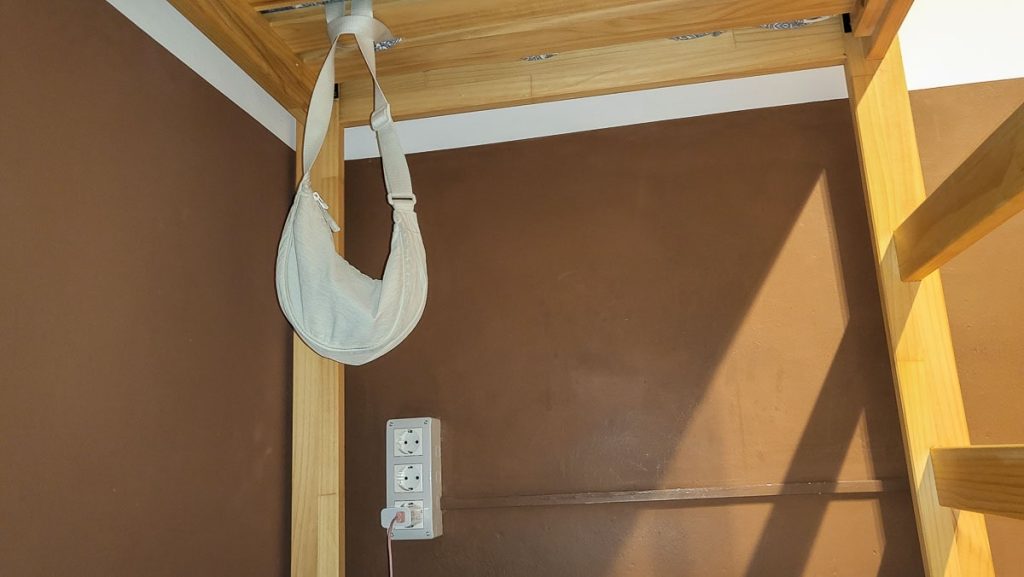 A small beige shoulder bag hanging from the wooden frame of a bunk bed in a hostel. Below the bag, there is a set of electrical outlets on the brown wall, with a phone charger plugged in. The scene is lit by warm lighting, casting soft shadows.