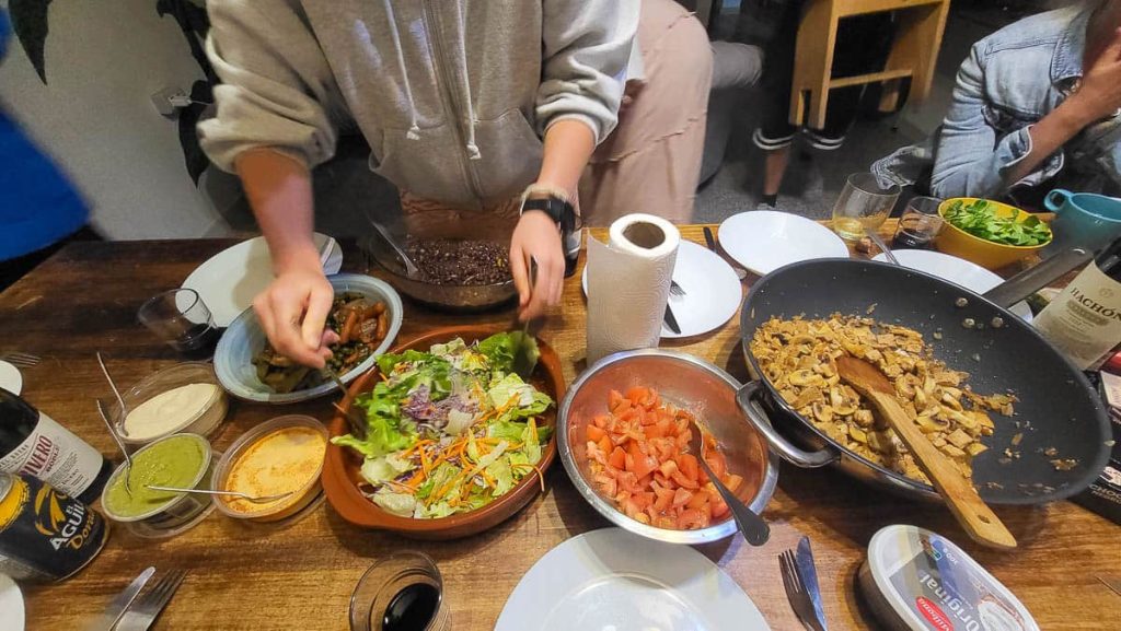 A wooden table filled with homemade dishes including a fresh salad, diced tomatoes, sautéed mushrooms, and various dips. A person in a grey hoodie is preparing their plate, while others are seated around the table, enjoying the meal.