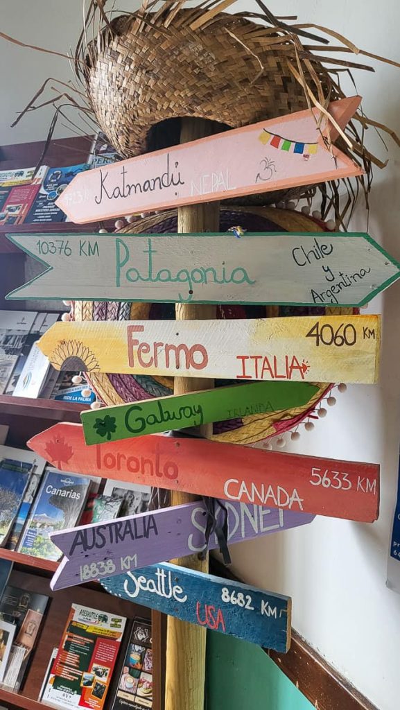 A wooden signpost with colorful arrows pointing in different directions, indicating distances to various destinations around the world. Locations include Kathmandu, Patagonia, Fermo, Galway, Toronto, Sydney, and Seattle, with handwritten text and small illustrations on each sign. A woven straw hat sits atop the post, and travel brochures are displayed in the background.