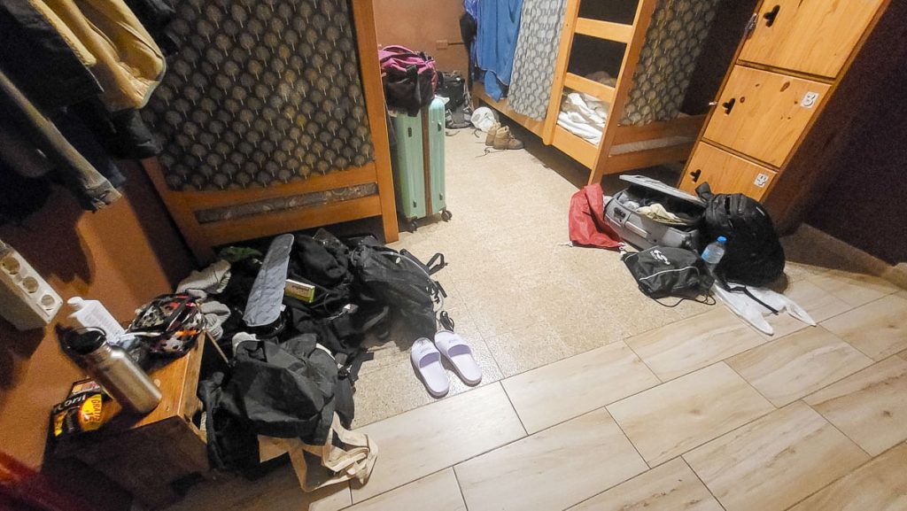 A cluttered hostel room with wooden bunk beds, backpacks, open suitcases, and scattered belongings covering the floor. A pair of slippers, a reusable water bottle, and various clothing items are visible. The room has a mix of brown walls and tiled flooring, with personal items hanging from the bunk beds.