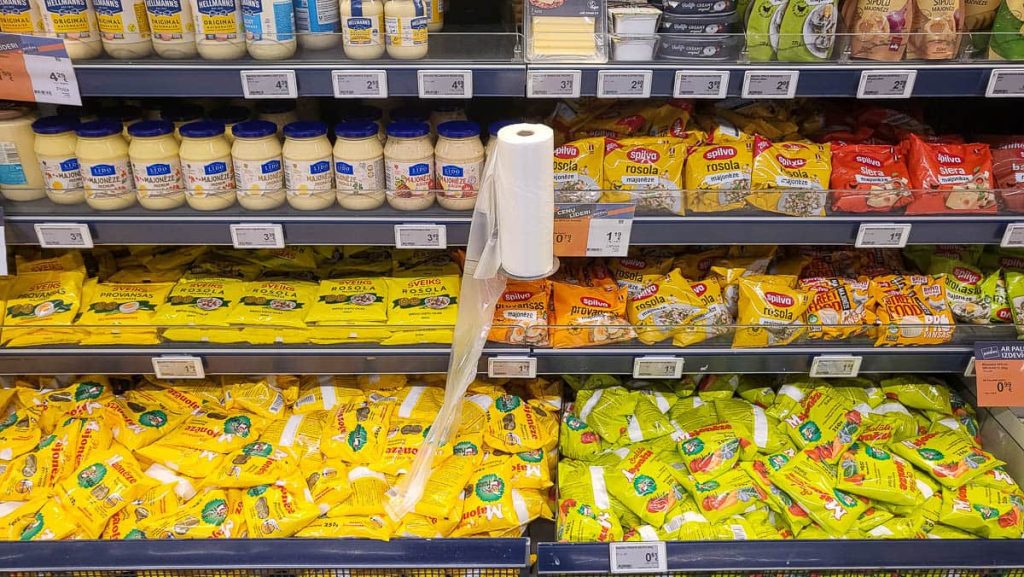 A supermarket shelf stocked with various brands of mayonnaise and salad dressings in jars and plastic pouches. The yellow packaging dominates the lower shelves, with some red and green packages mixed in. A roll of plastic bags for weighing produce hangs in the middle of the display.