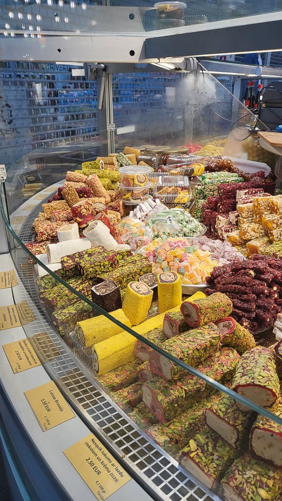 A close-up of a sweets display at Riga Central Market, with a variety of brightly colored Turkish delights, nougats, and pistachio-covered sweets arranged in a glass case. Small price tags in euros are visible, indicating the cost per item.