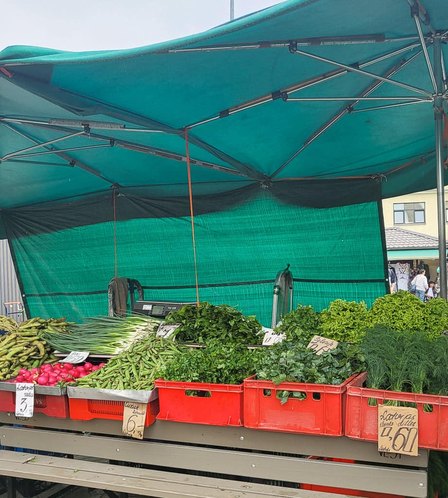 A vegetable stall at Riga Central Market covered by a green canopy, displaying fresh greens, herbs, radishes, scallions, and pea pods in red crates. Handwritten signs with prices in euros are placed among the produce.